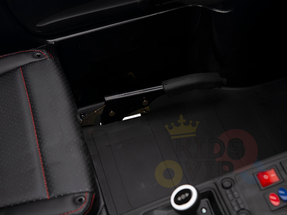 Close-up of the White 24V XXL SuperSport Ride On Car interior featuring a black grip handbrake next to a black padded seat with red stitching; floor has a rubber mat and console buttons are partially visible.