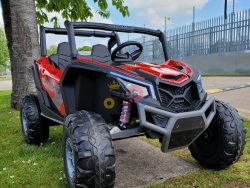 A Spyder Red XXL 2 Seats Challenger 24V MX Buggy MP4 Edition 4x4 Kids Ride On Car/UTV with RC and rubber wheels is parked on the grass near a tree. A clear plastic bottle sits inside. In the background, a metal fence stretches under the expansive blue sky.