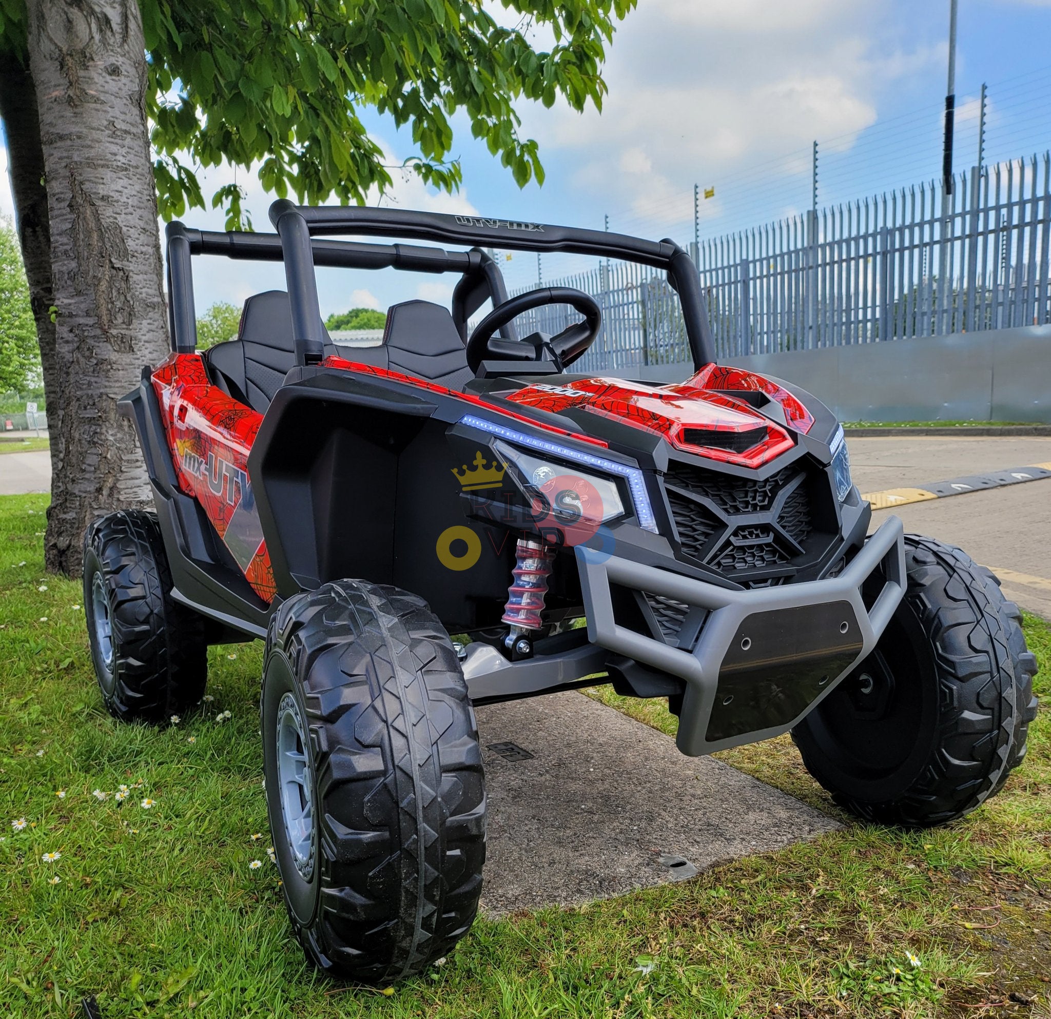 A Spyder Red XXL 2 Seats Challenger 24V MX Buggy MP4 Edition 4x4 Kids Ride On Car/UTV with RC and rubber wheels is parked on the grass near a tree. A clear plastic bottle sits inside. In the background, a metal fence stretches under the expansive blue sky.