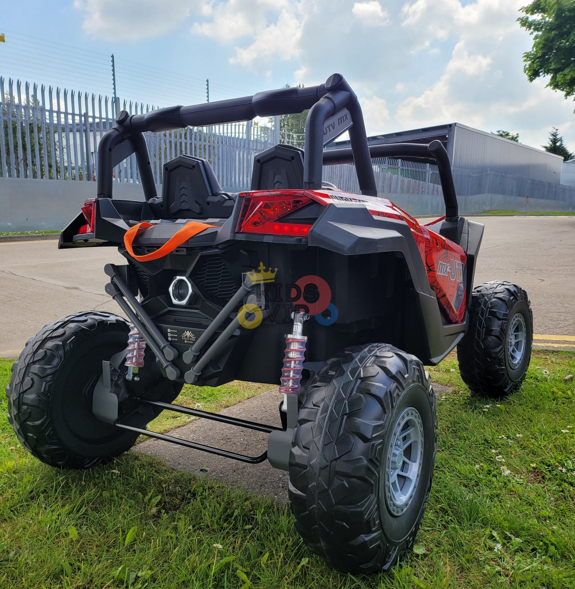 A Spyder Red XXL 2 Seats Challenger 24V MX Buggy MP4 Edition 4x4 Kids Ride On Car sits on the grass near a paved area under a partly cloudy sky. A tall metal fence and trees create an exciting off-road vibe for little explorers.