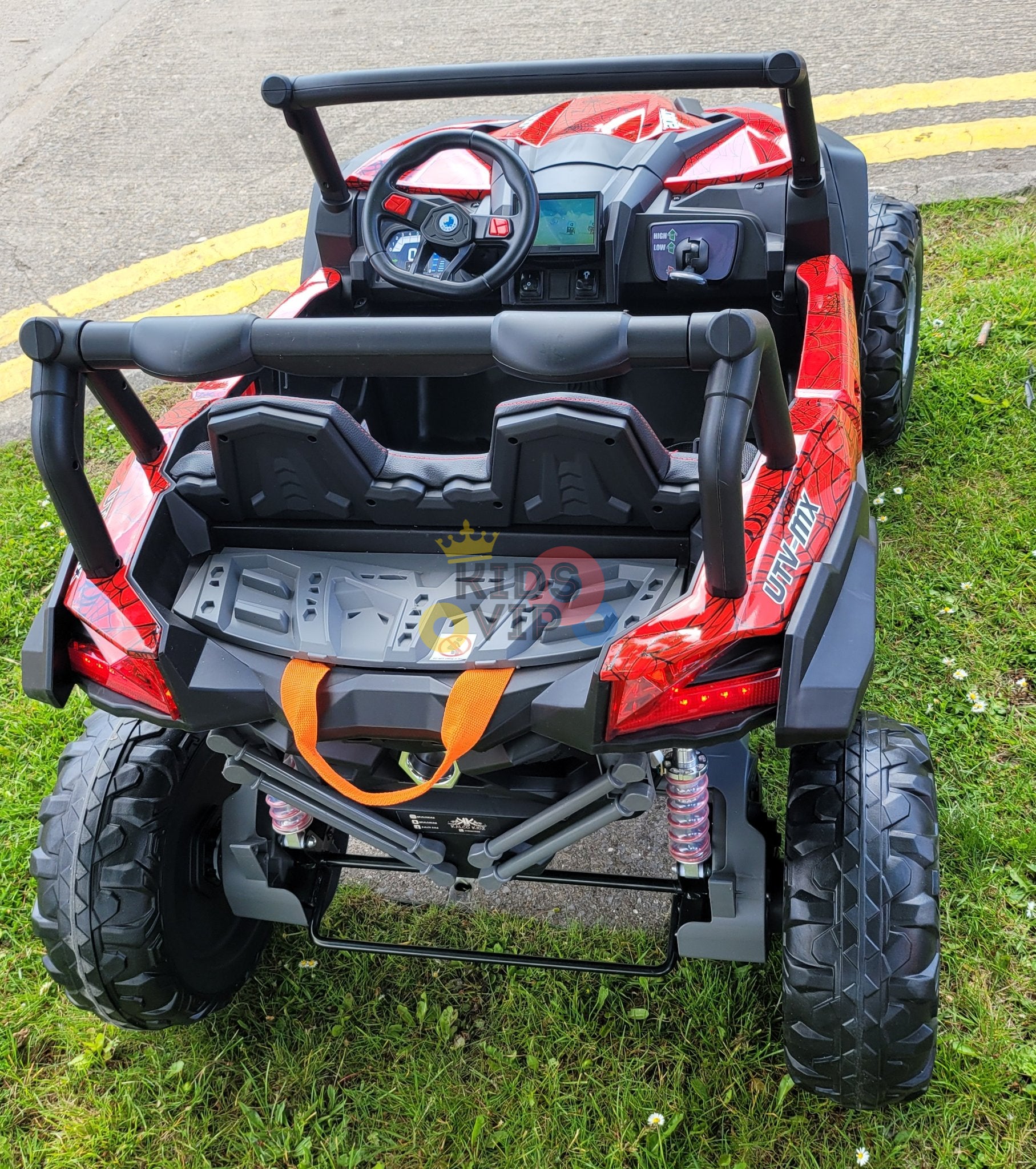 A Spyder Red XXL 2 Seats Challenger 24V MX Buggy MP4 Edition 4x4 Kids Ride On Car with RC and rubber wheels sits on the grass by a sidewalk. Resembling an off-road vehicle, it features a steering wheel, seatbelts, and dashboard for thrilling young explorers.
