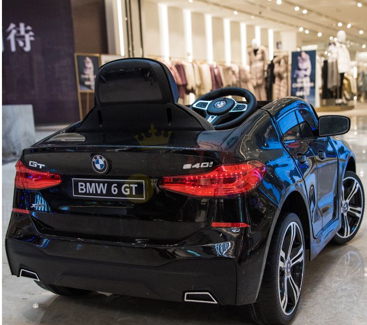 A Black Licensed BMW GT 12V Kids Ride On Power Car with Remote Control is parked indoors in front of a clothing store, with mannequins and racks visible. The ride-on toy boasts shiny details and a steering wheel, resembling a miniature luxury vehicle with upgraded features.