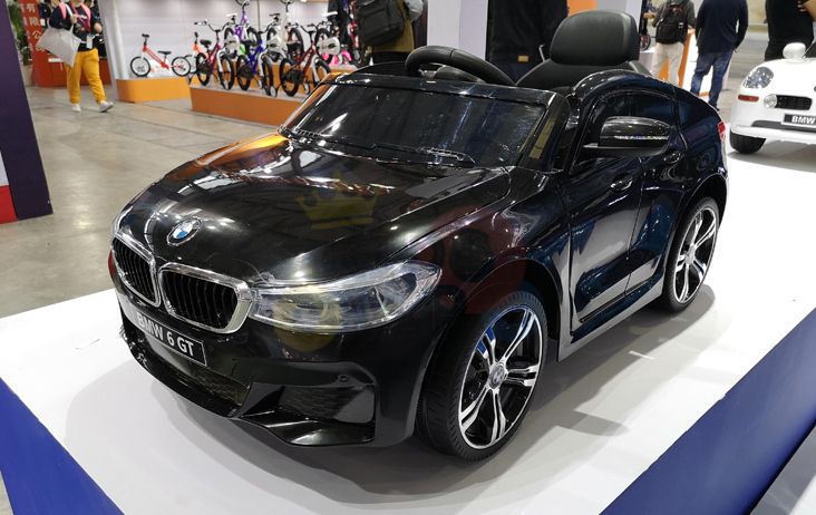 This stylish Black Licensed BMW GT 12V Ride On Power Car for kids, complete with remote control, is showcased indoors on a white platform. Its sleek design captivates, standing out amidst the backdrop of people and bikes to embody luxury in miniature form.