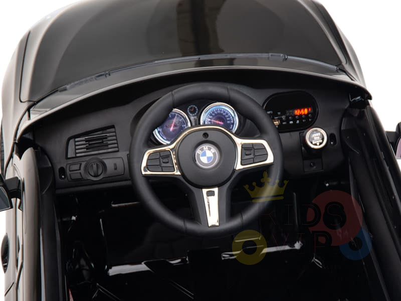 Close-up of a Black Licensed BMW GT 12V Kids Ride On Car dashboard with BMW logo on the steering wheel. Features include two circular gauges, a digital display on the right, buttons, and air vents on the left in this official electric ride-on.