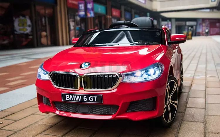 A Red 12V BMW GT Kids Licensed 1 Seater Ride On Car with Rubber Wheels and Remote is parked on a brick path. It features shining headlights and detailed design, resembling the real car, with blurred storefronts in the background enhancing the vibrant scene.