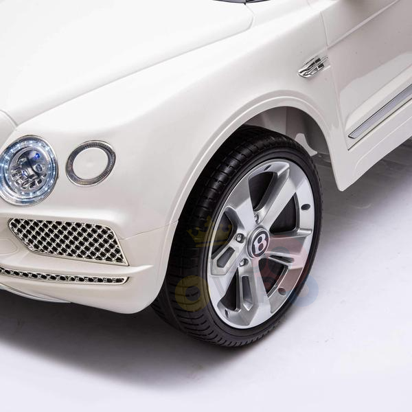 The front of the White 12V Bentley Bentayga Kids Licensed Ride On Car showcases a headlight, grille, and shiny rimmed wheel with a logo. It features a sleek, modern design and is perfectly sized for young drivers to enjoy their adventures.