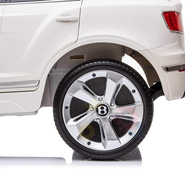 Close-up of a Bentley Bentayga Ride On Cars rear section displays its white body and detailed black and silver wheel with a stylized B center, reminiscent of the White 12V Bentley Bentayga Kids Licensed 1 Seater Ride On Car with Rubber Wheels, Remote, on a white reflective surface.