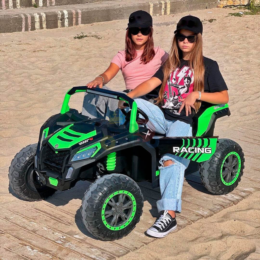Two young people enjoy their thrilling adventure on sandy terrain in a green and black 2 Seater XXL Blade BT 4WD Edition 24V Kids Ride On UTV, Buggy with RC, labeled RACING, featuring large wheels for an off-road experience. Theyre dressed casually and wear sunglasses.