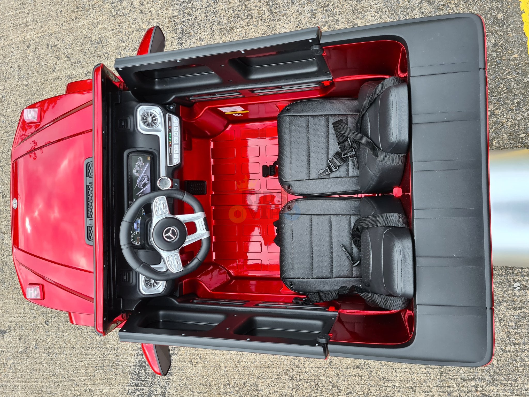 Aerial view of the red 2 Seats 4x4 Official Mercedes Benz G63 Complete Edition 24V Kids Ride On Car with RC, featuring two black seats, a steering wheel, and a basic dashboard. This sleek ride-on car is parked on a textured outdoor surface.