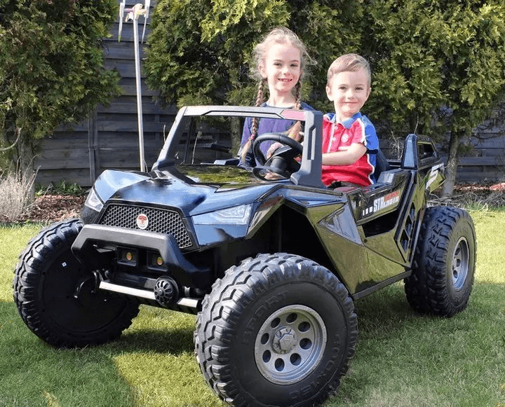 Two kids in a Red 24V/4X4 Dune Buggy Ride On UTV sit on grass. The long-haired girl, smiling, is the driver. Her friend in a colorful shirt sits next to her. Greenery and a wooden fence are in view, with a parental RC ensuring safety.