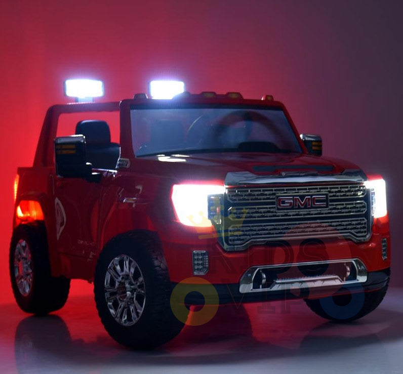 The blue 2-seat 4WD GMC Sierra Kids Ride On Car, officially licensed and powered by dual 12V batteries, features gleaming headlights, two small roof lights, a detailed grille, side mirrors, large shiny wheels, and plush leather seats. The illuminated backdrop enhances the trucks bright lights.