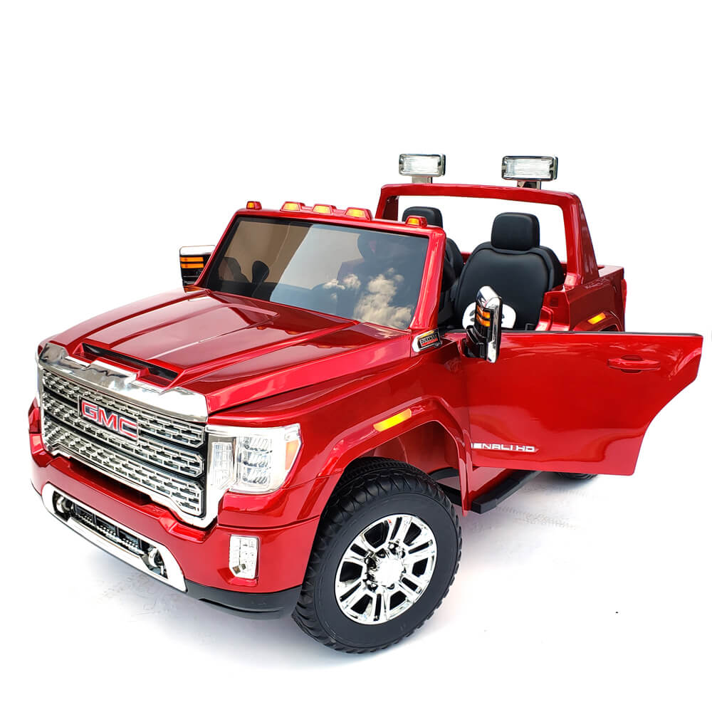 The 12V/2 seater ride-on car, styled like a GMC Sierra Denali, has a red body with an open drivers side door featuring silver accents. It includes a detailed grille, large wheels, extra roof and bumper lights, and a black interior with a visible steering wheel.