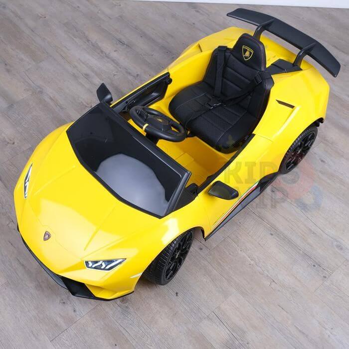A Yellow Officially Licensed 12V Lamborghini Huracan Sports Edition Ride on Car with black details, a spoiler, realistic features like a black steering wheel, remote control capabilities, and leather seats for children is parked on a wooden floor.