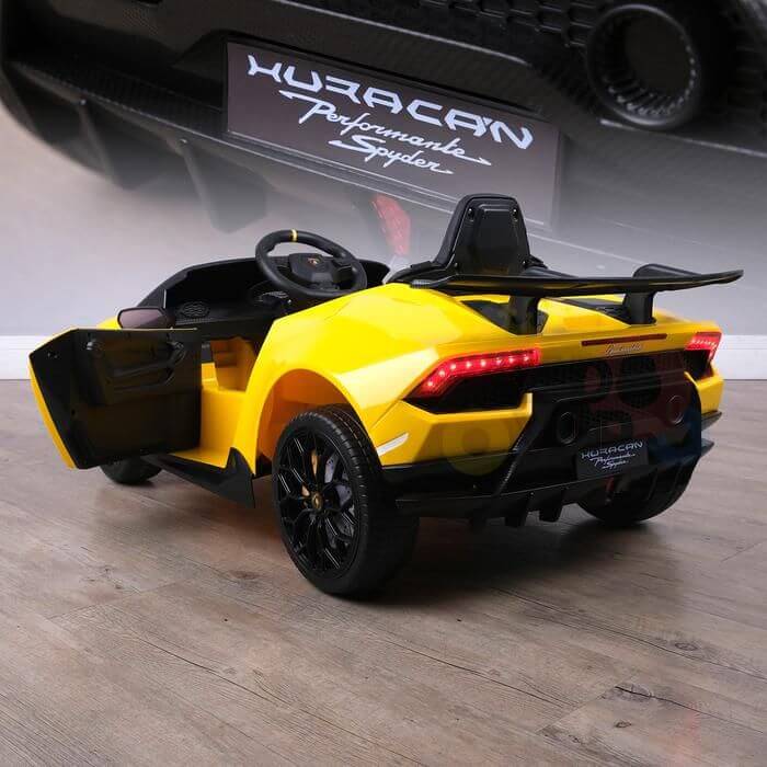 A Yellow Officially Licensed 12V Lamborghini Huracan Sports Edition Ride on Car, resembling a Huracán Performante Spyder with an open door, sits on a wooden floor. It features a steering wheel and remote control, rear lights visible, with its blurred logo in the background.