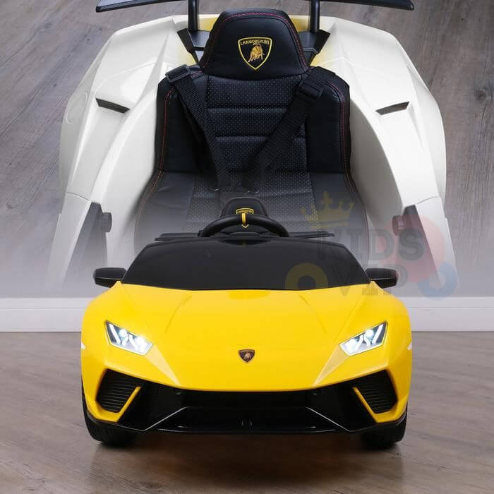 A Yellow Officially Licensed 12V Lamborghini Huracan Sports Edition Ride-On Car, with headlights on, is positioned before a larger white seat. The leather seat features black leather and red stitching with the iconic logo and rests elegantly on a wooden floor.