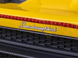 A close-up of the Officially Licensed 12V Lamborghini Huracan Sports Edition Ride-on Car reveals a yellow panel and hexagonal mesh grille, highlighted by the silver cursive Lamborghini logo. In the background, a part of the black leather seat is visible inside.