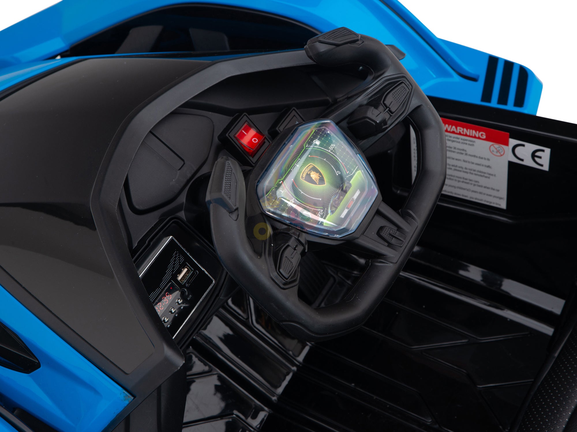 Close-up of the black steering wheel with a multi-colored emblem, prominent dashboard, and control buttons of the Blue Official Complete Edition 12V Lamborghini Vision GT Kids Ride-On Car, featuring a mainly black interior with an Eco Leather Seat and visible side warning label.