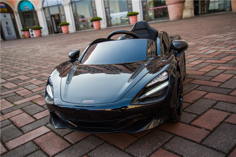 A Black 12V McLaren 720S Kids Licensed 1 Seater Ride On Car with Rubber Wheels and Remote is parked on patterned brick pavement. The sleek toy features detailed headlights, a glossy finish, and upgraded features, with shops and large potted plants in the background.
