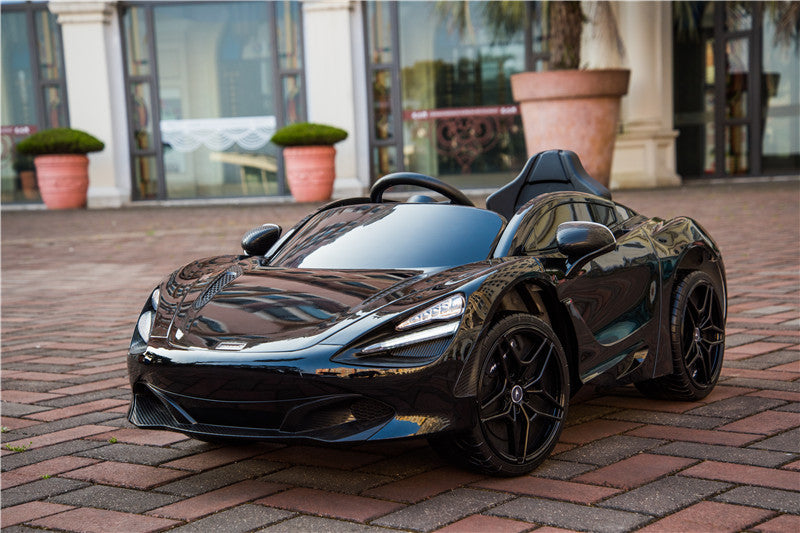 A shiny, Black 12V McLaren 720S Kids Licensed 1 Seater Ride-On Car with rubber wheels and a remote is parked on brick pavement, showcasing sleek curves and a convertible style. In the background are large windows and potted plants.