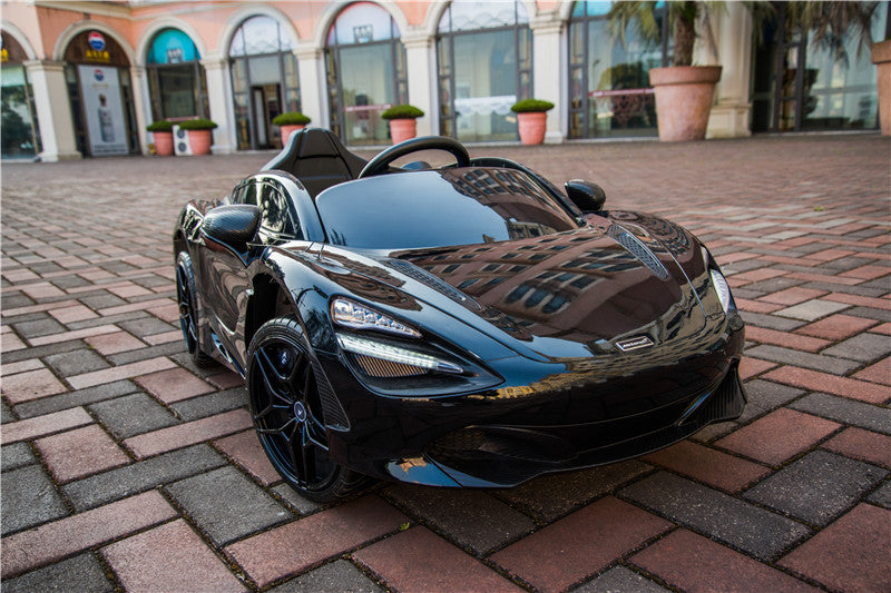 A sleek Black 12V McLaren 720S Kids Licensed 1 Seater Ride On Car with Rubber Wheels and Remote is parked on a brick pavement, set against a backdrop of an arched building, large potted plants, and shop windows highlighting this upgraded remote control supercar.