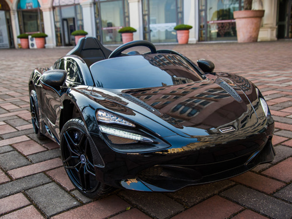 A Black 12V McLaren 720S Kids Licensed 1 Seater Ride On Car with rubber wheels is parked on a brick-paved area. The stylish remote control car is set against a backdrop of shopfronts with large windows and potted plants, highlighting its sporty allure in an urban environment.