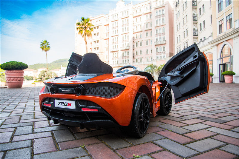 An Orange 12V McLaren 720S Kids Licensed Ride On Car with butterfly doors is parked on a cobblestone street before tall, elegant buildings. Palm trees and mountains appear in the background beneath a partly cloudy sky, offering the ultimate Ride On Super Car adventure.