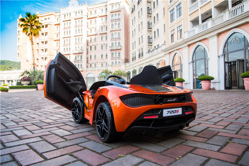 An Orange 12V McLaren 720S Kids Licensed 1 Seater Ride On Car with rubber wheels and remote, designed to resemble a sleek McLaren, is parked in a courtyard. The background showcases a large elegant building with columns and arches.