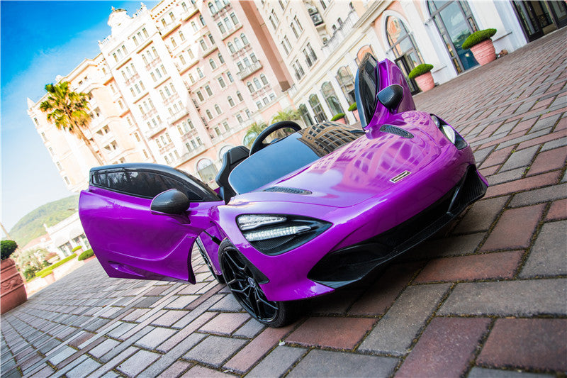 A purple 12V McLaren 720S kids licensed ride-on car with rubber wheels and remote, doors open, is parked on a brick pavement. Tall buildings and a palm tree stand in the background under a clear blue sky.