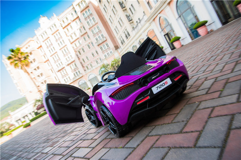 A Purple 12V McLaren 720S Kids Licensed 1 Seater Ride On Car with rubber wheels and open doors is parked on a brick pathway in front of tall buildings, with palm trees and hills under a clear blue sky.