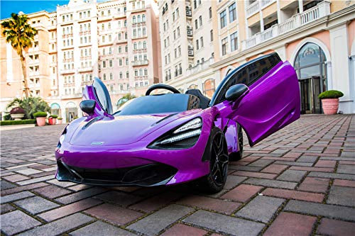 A sleek Purple 12V McLaren 720S Kids Licensed Ride On Car with butterfly doors glistens on a cobblestone path under the clear sky. Surrounded by palm trees and plants, it sparkles majestically before a grand building, capturing every childs dream of adventure.