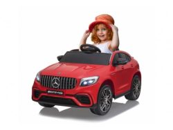 A young red-haired child beams while sitting in a ride-on Mercedes-Benz AMG with glowing headlights. Featuring the iconic logo and AMG branding, this toy includes the Enhance Your Driving Experience with a 12V Mercedes GLC Steering Wheel Upgrade, perfect for their driving adventure.