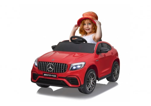 A child with red hair and a red hat smiles while sitting in the 12V Mercedes GLC ride-on car, which features headlights, the iconic logo, and black wheels with silver accents.