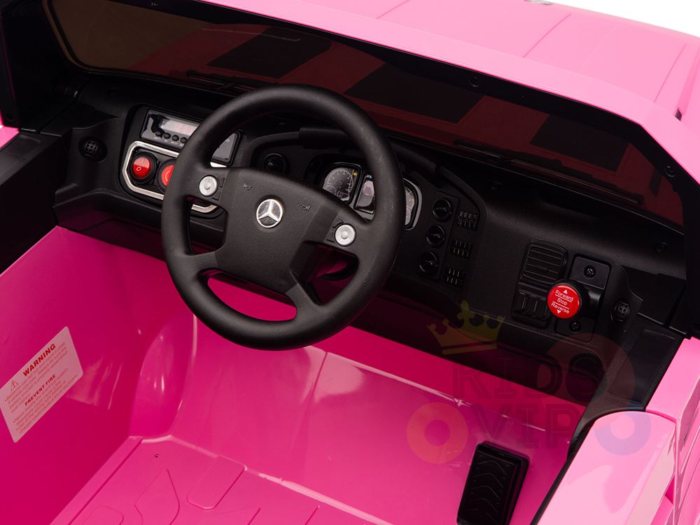 The Pink 12V Mercedes Benz Zetros Kids Ride On Truck features a modern interior with a pink dashboard, black steering wheel with silver logo, playful design elements, various controls and buttons, all powered by a 12V12amp system.