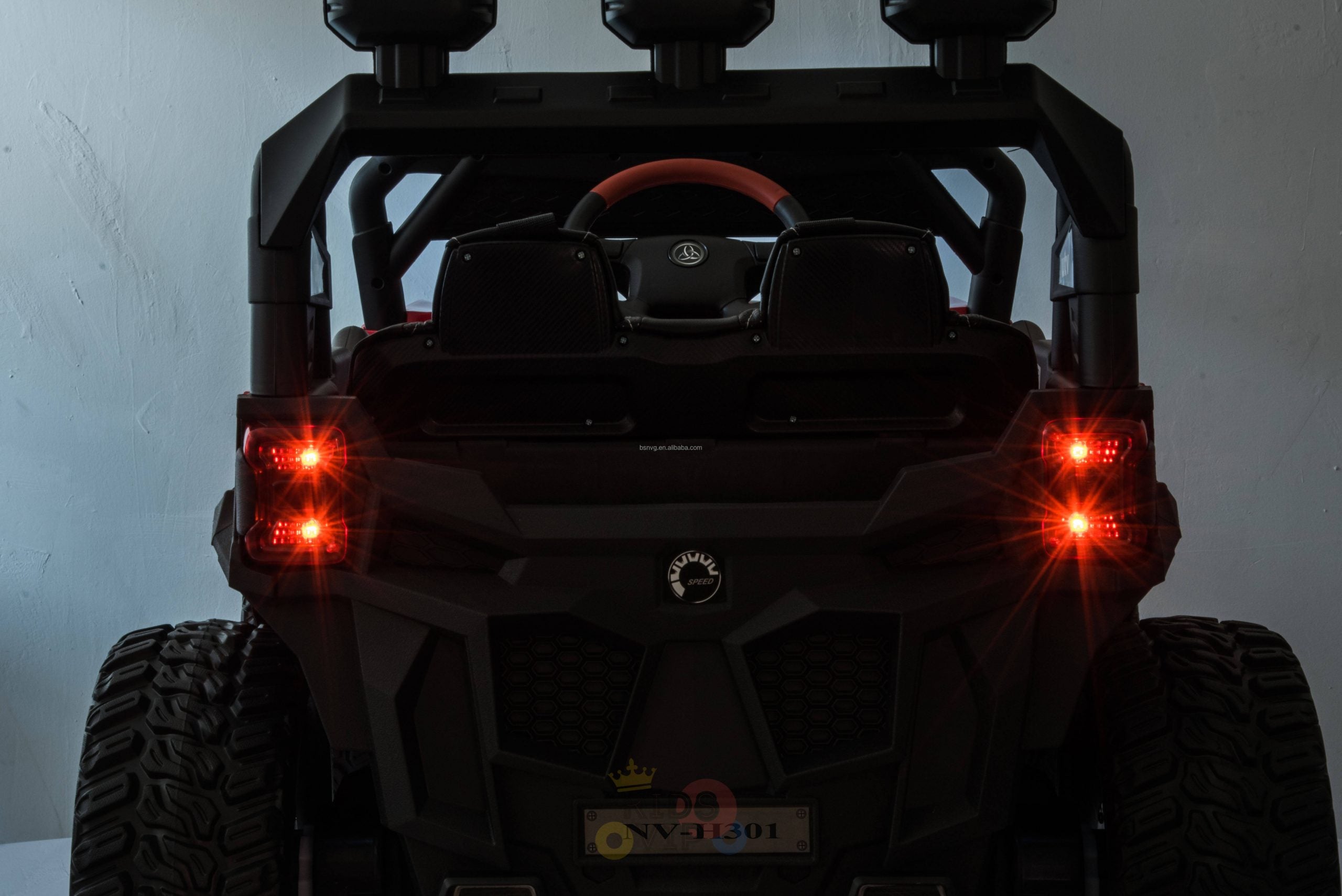 The image shows the rear view of the Upgraded 12V Junior Sport Utility Ride On Buggy/UTV 4WD Edition, featuring large tires and red lights. It highlights a sleek, rugged off-road design with a small license plate and steering wheel, set against a plain wall backdrop.